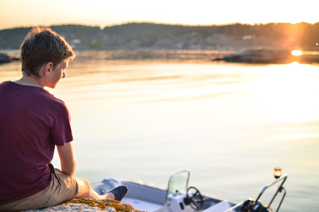 A boy sits on a dock beside a small boat, enjoying the tranquil sunset over a calm lake.